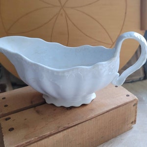 May include: A white ceramic gravy boat with a fluted design and a handle. The boat is sitting on a wooden surface.