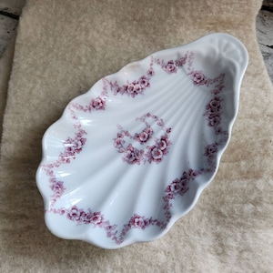 May include: A white porcelain shell-shaped dish with a pink floral design. The dish is perfect for serving small snacks or holding jewelry.