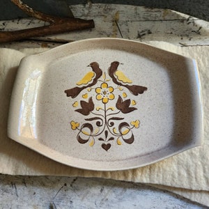 May include: A beige ceramic serving tray with a brown and yellow floral design featuring two birds. The tray has a speckled finish and a slightly rounded rectangular shape.