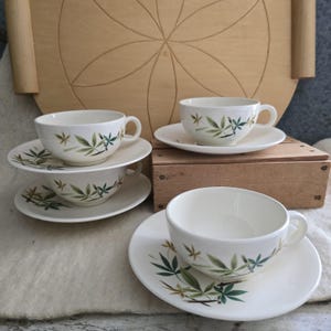 May include: A set of four white teacups and saucers. Each cup and saucer features a green and brown leaf design. The cups are stacked on top of each other, and the saucers are arranged around them. The teacups are on a white surface.