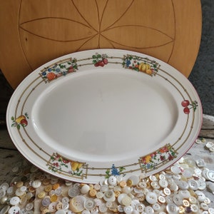 May include: A white oval serving platter with a red rim and a gold and green floral design featuring fruits and vegetables. The design includes a pumpkin, grapes, apples, and pears.