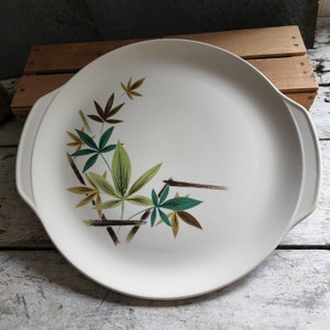 May include: A white ceramic serving platter with two handles. The platter features a stylized design of green and brown leaves with brown branches.