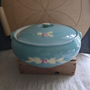 May include: A teal ceramic casserole dish with a lid. The dish features floral accents in pink and yellow. The round dish sits on a small wooden crate. The lid has a small knob for easy handling.
