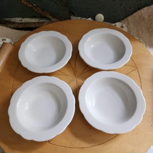 May include: A set of four white ceramic bowls, each featuring a smooth, glossy finish and a distinct scalloped edge design. The bowls are circular in shape with a deep center and a wide, curved rim, presenting a clean and uniform appearance.