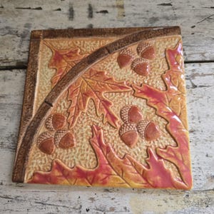 May include: A square ceramic tile featuring a raised relief design of autumn leaves and clusters of acorns. The surface has a textured background with a color palette of warm yellow, orange, and deep red, accented by a dark brown curved border.