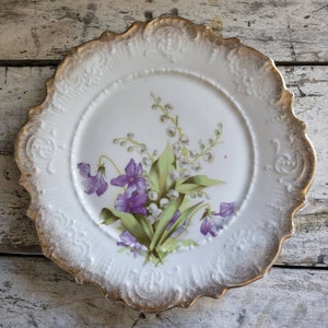 May include: White porcelain plate with a gold rim and floral design. The plate features a bouquet of purple violets, white lily of the valley, and green leaves. The rim has a scalloped edge with embossed detailing.
