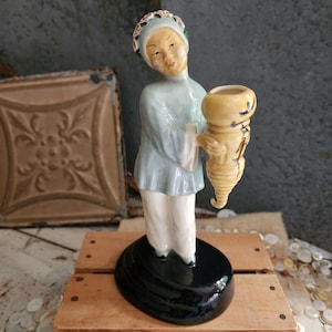May include: A vintage porcelain figurine of a woman in traditional attire, holding a yellow vase. The figurine has a pale blue robe, white pants, and a floral headpiece. It stands on a black, glossy base. The background includes a wooden surface and a decorative metal panel.