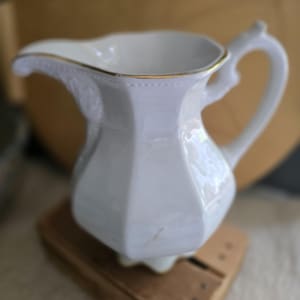 May include: A white ceramic creamer pitcher featuring a faceted, hexagonal body design, a curved handle, and a wide pouring spout. The upper rim of the pitcher is accented with a thin, metallic gold trim, providing a subtle contrast to the glossy white finish.