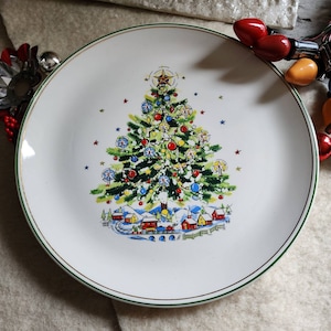 May include: A white ceramic Christmas plate with a green rim and a gold trim. The plate features a colorful illustration of a Christmas tree with ornaments, a star on top, and a small village at the base.