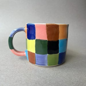 May include: A ceramic mug with a colourful checkerboard pattern. The mug has a white interior and a curved handle. The squares are painted in various colours, including blue, pink, yellow, green, and brown. The mug is a unique piece of kitchenware.