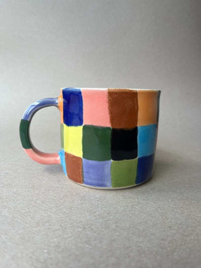 May include: A ceramic mug with a colorful checkerboard pattern. The mug features a white interior and a curved handle. The squares are painted in various colors, including blue, pink, yellow, green, and brown. The mug is a unique piece of kitchenware.