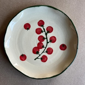 May include: A cream-coloured ceramic plate with a green rim. The plate features a design of red tomatoes, with a cluster and individual tomatoes scattered across the surface.