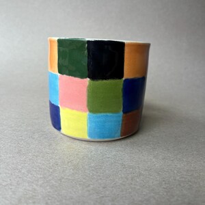May include: A ceramic planter with a colorful checkerboard pattern. The squares are painted in various colors, including orange, green, black, pink, yellow, blue, and brown. The planter has a cylindrical shape and a white interior.