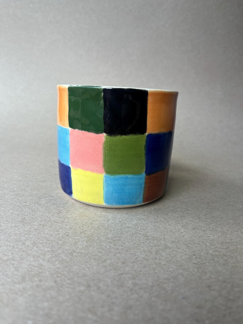 May include: A ceramic planter with a colorful checkerboard pattern. The squares are painted in various colors, including orange, green, black, pink, yellow, blue, and brown. The planter has a cylindrical shape and a white interior.
