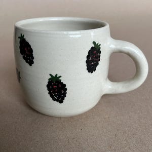 May include: A cream-colored ceramic mug with a rounded body and a handle. The mug is decorated with hand-painted black and green blackberries. The mug is suitable for hot or cold beverages.