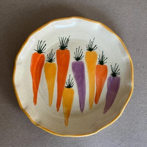 Może przedstawiać: Ceramiczny talerz z kremową podstawą i falistym, żółtym obramowaniem. Talerz zdobią ręcznie malowane marchewki w odcieniach pomarańczu, żółci i fioletu, każda z zielonymi wierzchołkami. Unikalny i kolorowy element do serwowania potraw.