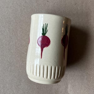 May include: A cream-coloured ceramic mug featuring a hand-painted design of red beetroot with green tops. The mug has a cylindrical shape with vertical grooves near the base. The design is repeated around the mug.