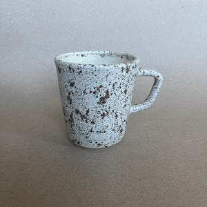 Op de afbeelding: Een wit keramieken mok met bruine spikkels en een oor. De mok heeft een uitlopende vorm. Het oor is een simpele lus. Het gespikkelde patroon geeft een rustieke uitstraling. Geschikt voor thee of koffie, de mok lijkt handgemaakt.