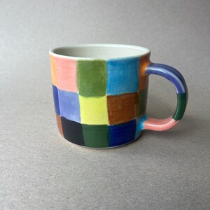 May include: A ceramic mug with a colorful checkerboard pattern. The squares are in shades of pink, green, blue, yellow, brown, and black. The handle is a gradient of colors, and the interior is white.