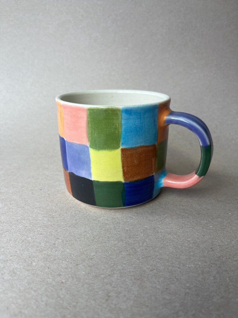 May include: A ceramic mug with a colorful checkerboard pattern. The squares are in shades of pink, green, blue, yellow, brown, and black. The handle is a gradient of colors, and the interior is white.