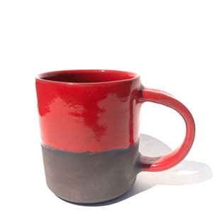 Puede incluir: Una taza de cerámica roja y marrón con un asa redondeada. La taza está esmaltada en rojo brillante en la mitad superior y en marrón mate en la mitad inferior.