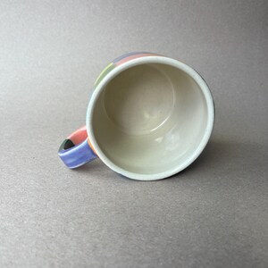 May include: A ceramic mug with a colorful, striped design around the rim and handle. The stripes include pink, green, blue, and yellow. The interior of the mug is white, and the handle is a mix of colors.