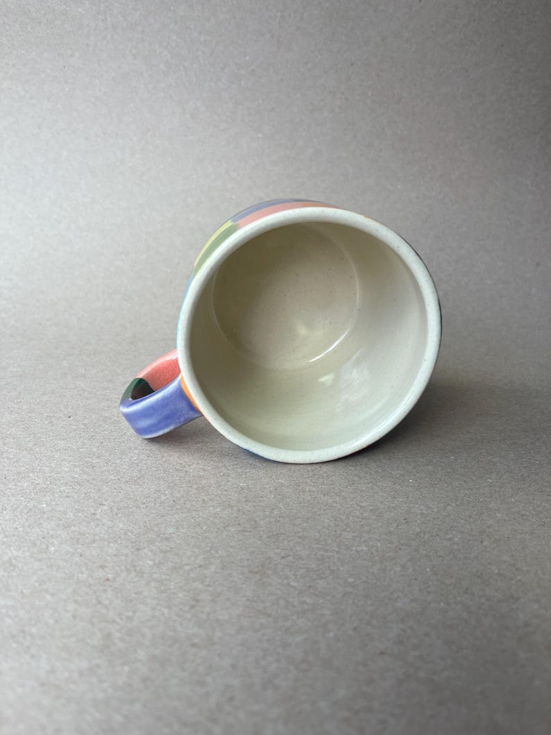 May include: A ceramic mug with a colorful, striped design around the rim and handle. The stripes include pink, green, blue, and yellow. The interior of the mug is white, and the handle is a mix of colors.