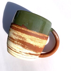 May include: Handmade ceramic mug with a green top and a brown and cream-coloured striped design. The mug has a rounded brown handle and is set against a white background. A shadow is cast on the white surface.