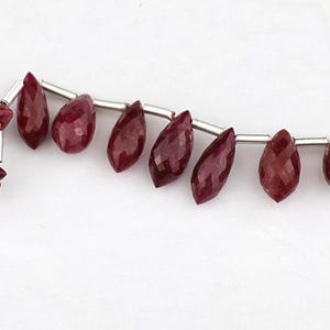 May include: A strand of faceted, deep red, teardrop-shaped gemstone beads alternating with small, cylindrical silver-toned metal spacers. The beads are strung on a thin, reddish-brown thread that extends slightly from both ends of the arrangement.