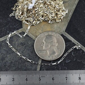 May include: A silver-colored metal chain with rectangular links, shown next to a United States quarter coin and a metal ruler that displays measurements in millimeters and centimeters to provide a clear sense of scale for the chain links.
