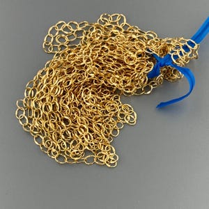 May include: A long, continuous gold-colored metal chain composed of numerous small, interlocking oval links. The chain is gathered in a loose pile and is secured at one end by a bright blue plastic zip tie that holds the links together.