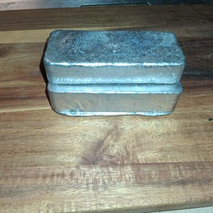 May include: Two silver-coloured metal bars, possibly ingots, are stacked on a wooden surface. The metal has a slightly textured appearance. The wooden surface displays a prominent grain pattern. The items are rectangular in shape.