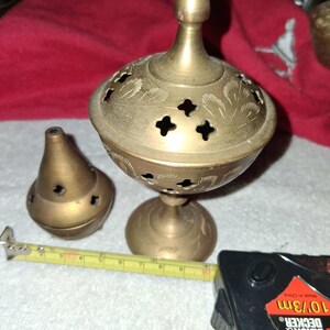 May include: A pair of vintage brass incense burners. The larger burner has a domed lid with floral cutouts and a pedestal base. The smaller burner is cone-shaped. A measuring tape is next to the burners.