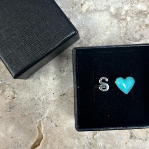 May include: A small black box with a turquoise heart and the letter "S" inside. The box is open and the heart and letter are visible.
