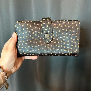 May include: A black and tan polka dot wallet with a button closure. The wallet is rectangular and has a flap with a metal button. The polka dots are tan and evenly spaced across the black background.