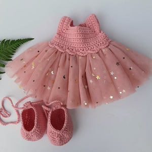 May include: A pink crocheted dress with a tulle skirt decorated with gold stars. The dress is paired with matching pink crocheted ballet shoes.
