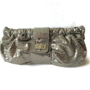 Può includere: Una pochette color argento metallizzato con una texture a pelle di serpente. La borsa rettangolare presenta un design arricciato e una patta centrale verticale chiusa da un meccanismo di bloccaggio a rotazione in metallo argentato.