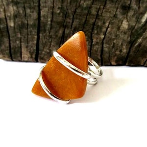 May include: A ring featuring a triangular, smooth, mustard-yellow stone held in place by a silver-colored metal wire that wraps around the stone and forms the band. The stone has a matte texture and a solid, opaque, warm yellow-orange color.