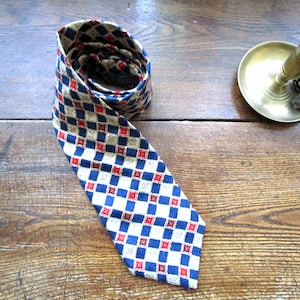 Puede incluir: Una corbata con un patrón repetitivo de diamantes y cuadrados en tonos azul marino, crema y rojo. La tela tiene una apariencia texturizada con pequeños detalles intrincados de punto de cruz centrados en cada cuadrado, dispuestos en cuadrícula.