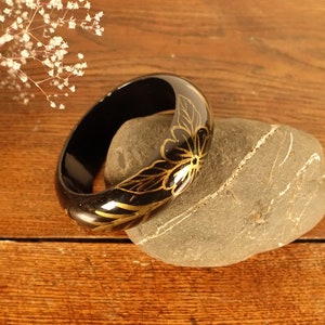 Könnte beinhalten: Ein glänzender schwarzer Armreif mit einem zarten goldfarbenen Blumenmuster aus Blättern und Blütenblättern. Das kreisförmige Accessoire hat eine glatte, reflektierende Oberfläche und ein abgerundetes Profil mit filigranen Linien in Gold.