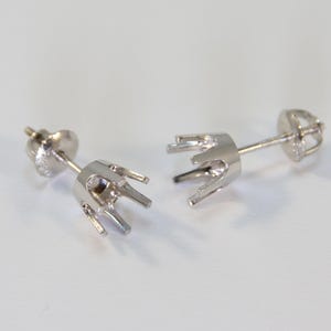 Può includere: Due orecchini a lobo color argento con un design a castone. Ogni orecchino presenta un perno e un'incastonatura decorativa per contenere una gemma. Gli orecchini sono esposti su una superficie bianca.