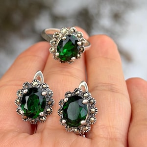 Puede incluir: Un conjunto de anillo y pendientes de plata. Cada pieza presenta una gran piedra preciosa ovalada de color verde esmeralda rodeada por un diseño floral. Las joyas se muestran sobre un fondo borroso y nevado.