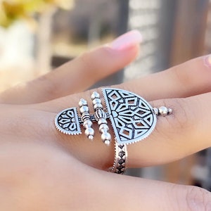 May include: A silver ring with a geometric design. The ring features a large, openwork pendant with a circular shape and intricate details. The pendant is attached to a thin band with a textured surface.
