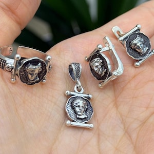 May include: A silver jewellery set consisting of a ring, a pendant, and a pair of earrings. Each piece features a circular medallion with a raised relief of a classical human face, framed by horizontal silver bars and a textured background.