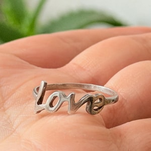 Op de afbeelding: Een zilveren ring met het woord "love" in cursief schrift. De ring wordt getoond op een hand, met een vage groene achtergrond. Het ontwerp van de ring heeft een delicaat, open lettertype, waardoor een romantische en elegante esthetiek ontstaat. De ring is een sieraad.