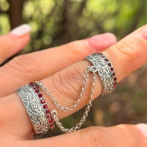 Pode incluir: Anel duplo prateado com detalhes florais ornamentados e pequenas pedras vermelhas. Os anéis são conectados por uma delicada corrente. O design se destaca em um fundo verde desfocado.