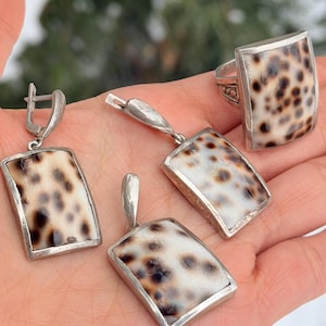 Könnte beinhalten: Ein Schmuckset mit einem Ring, Ohrringen und einem Anhänger. Jedes Stück hat ein rechteckiges Design mit einem silbernen Rahmen und einer gesprenkelten, cremefarbenen Mitte mit braunen Flecken. Das Set wird auf einer Hand präsentiert.