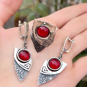 May include: A silver jewellery set comprising a ring and earrings, each with a large, round, red gemstone. The ring has a shield-like design, and the earrings are triangular. The metal has a textured, hammered appearance.