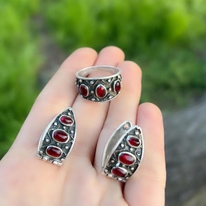 Op de afbeelding: Een set zilveren sieraden met rode edelsteen accenten. De set bevat een ring en een paar oorbellen. De ring heeft vijf edelstenen en de oorbellen hebben er elk vier.