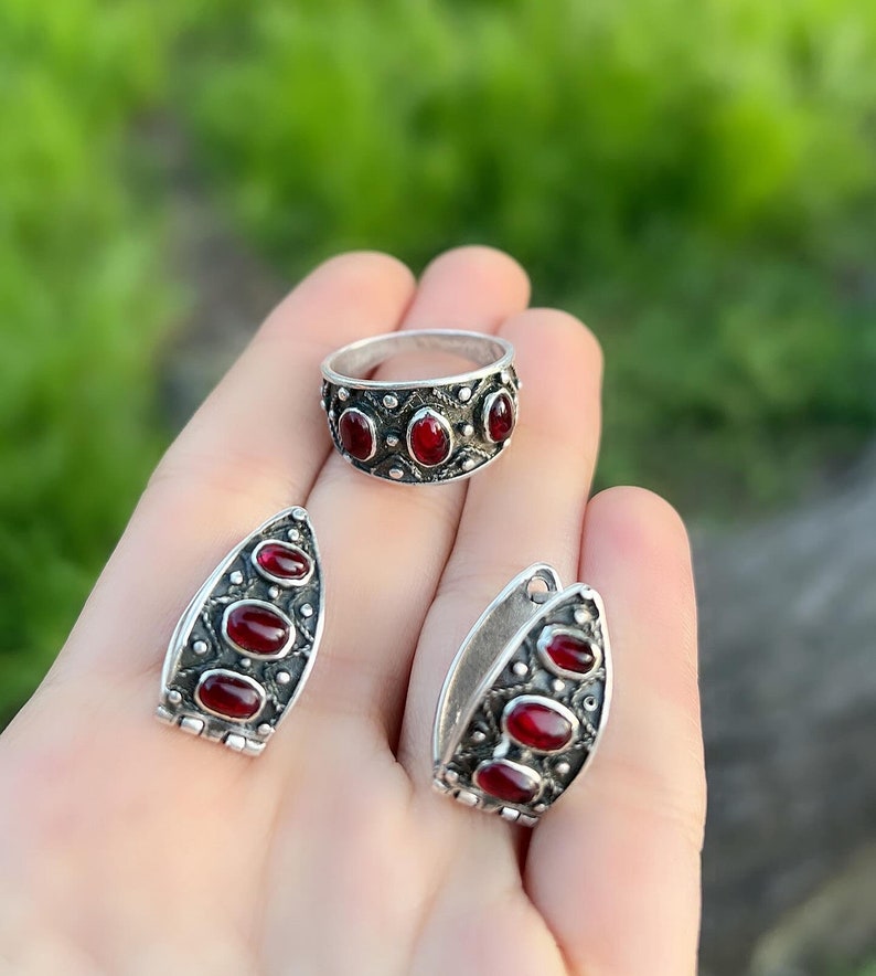 Op de afbeelding: Een set zilveren sieraden met rode edelsteen accenten. De set bevat een ring en een paar oorbellen. De ring heeft vijf edelstenen en de oorbellen hebben er elk vier.